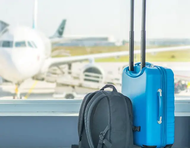 A blue suitcase and backpack sitting in front of an airport window.